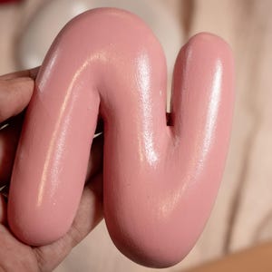 Puede incluir: Una escultura abstracta de madera rosa claro con forma de letra "N" curvada. La superficie lisa y brillante refleja la luz, resaltando las formas redondeadas. La escultura se sostiene en una mano, con un fondo neutro.