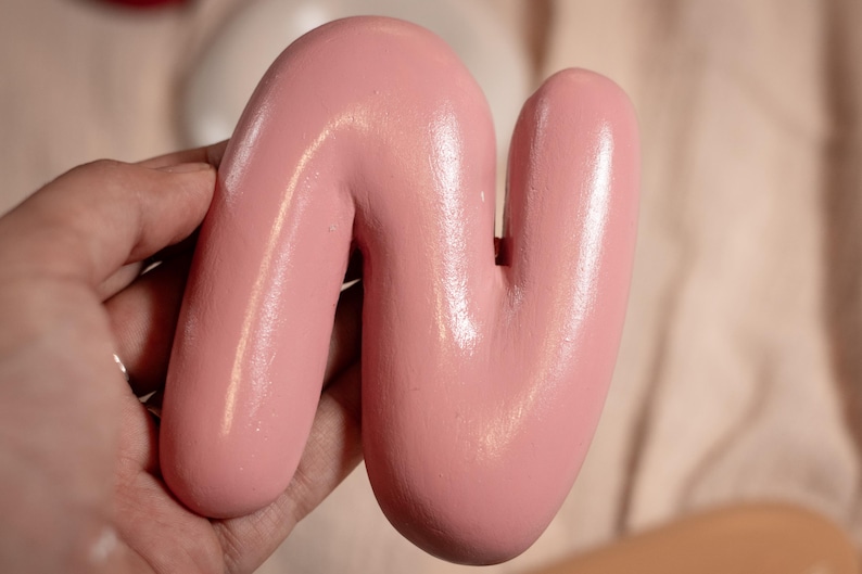 May include: A light pink, abstract wooden sculpture in the shape of a curved letter 'N'. The smooth, glossy surface reflects light. The sculpture is held in a hand against a soft, neutral background.