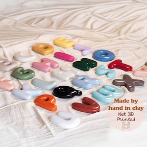 May include: A collection of individual, smooth, glossy, multi-colored clay letters of the alphabet arranged in a group. The text on the right side reads: Made by hand in clay, Not 3D Printed, with a small line drawing of hands forming a heart shape.
