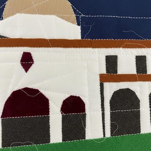 May include: A quilted textile depicting a building with a white facade, brown accents, and dark gray arches. The sky is navy blue, and the ground is green. The building has a dome and a diamond-shaped accent. The stitching is white.