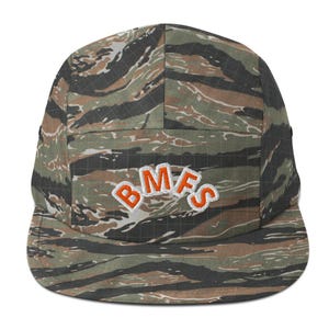 Könnte beinhalten: Eine Five-Panel-Cap mit einem Tigerstreifen-Tarnmuster in Grün-, Braun- und Schwarztönen. Auf der Vorderseite der Cap sind die Buchstaben "BMFS" in Orange mit weißer Umrandung gestickt.