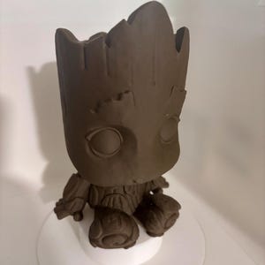 May include: A brown Groot figurine from Guardians of the Galaxy. The figure has large eyes and a textured body, seated on a white circular base. The figure is approximately 10 cm tall.