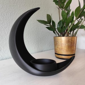 May include: A black, crescent moon-shaped candle holder. The holder has a circular space for a candle and is made of a matte black material. A gold-coloured pot with a green plant is in the background.