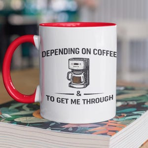 May include: White coffee mug with a red interior and handle. The mug features the text "DEPENDING ON COFFEE & TO GET ME THROUGH" along with a graphic of a coffee maker. The mug is sitting on a book.