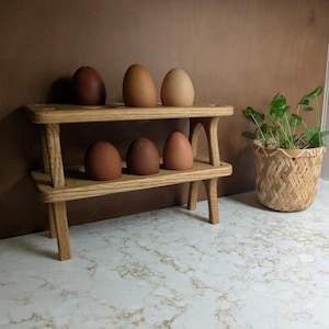 May include: A wooden two-tiered shelf displays six decorative eggs in varying shades of brown. A small woven basket holds a green plant. The shelf and eggs are on a white countertop with a brown wall in the background.