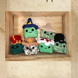May include: A wooden shelf displays seven crocheted figures, each with a different hat. The figures are green with black eyes and smiles. Hats include a witch's hat, a fedora, and a Santa hat. Text at the top reads "Hats & Hoppers."