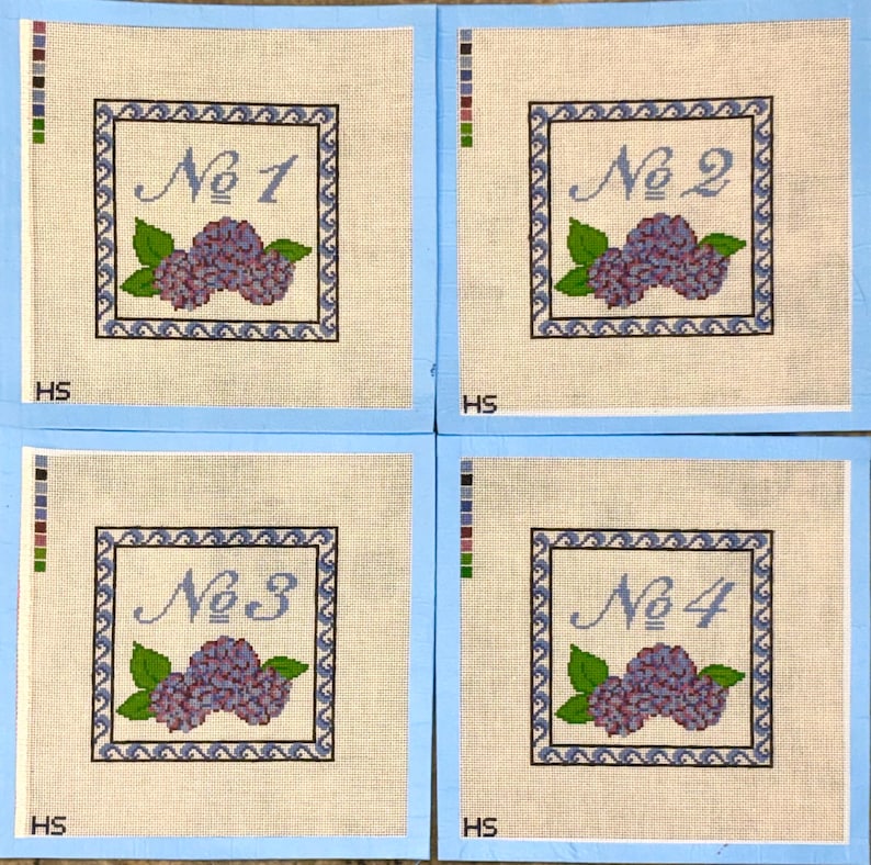 Op de afbeelding: Vier vierkante borduurkaders met lichtblauwe randen. Elk kader heeft een omlijnd ontwerp met een nummer, een tros paarse bloemen en groene bladeren. De nummers zijn "No 1", "No 2", "No 3" en "No 4".