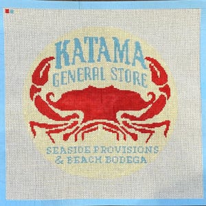 Op de afbeelding: Een borduurcanvas met een rood krabontwerp en de woorden "KATAMA GENERAL STORE" in blauw, en "SEASIDE PROVISIONS & BEACH BODEGA" in blauw. De achtergrond is gebroken wit met een gele gloed, en de rand is lichtblauw.