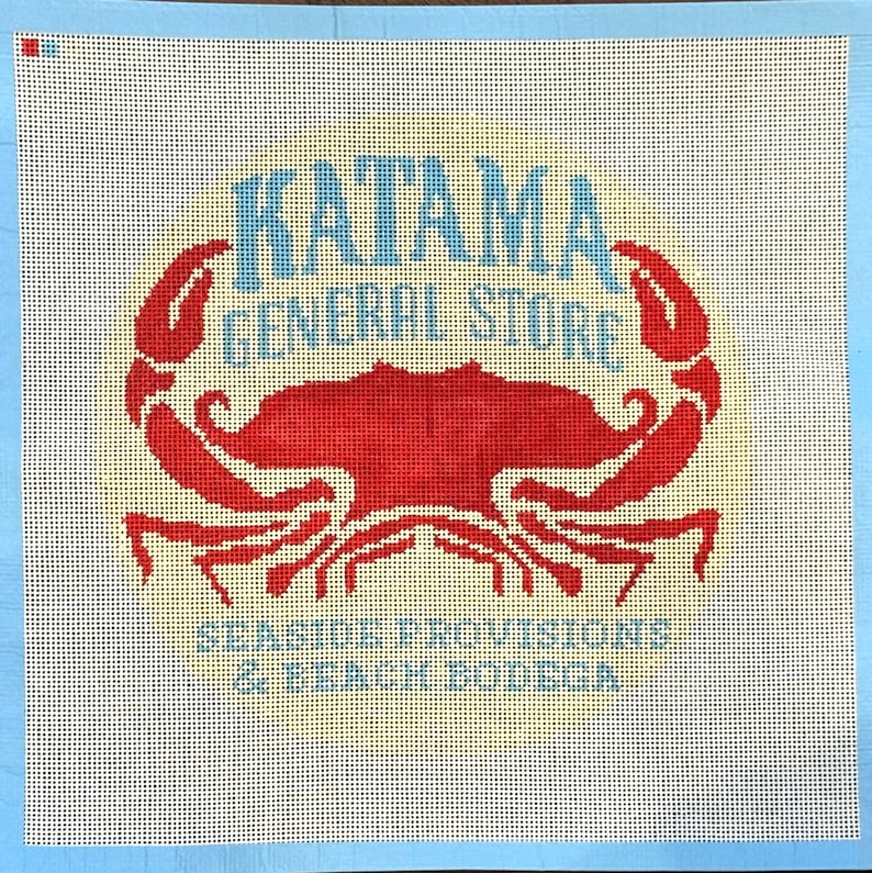 Op de afbeelding: Een borduurcanvas met een rood krabontwerp en de woorden "KATAMA GENERAL STORE" in blauw, en "SEASIDE PROVISIONS & BEACH BODEGA" in blauw. De achtergrond is gebroken wit met een gele gloed, en de rand is lichtblauw.