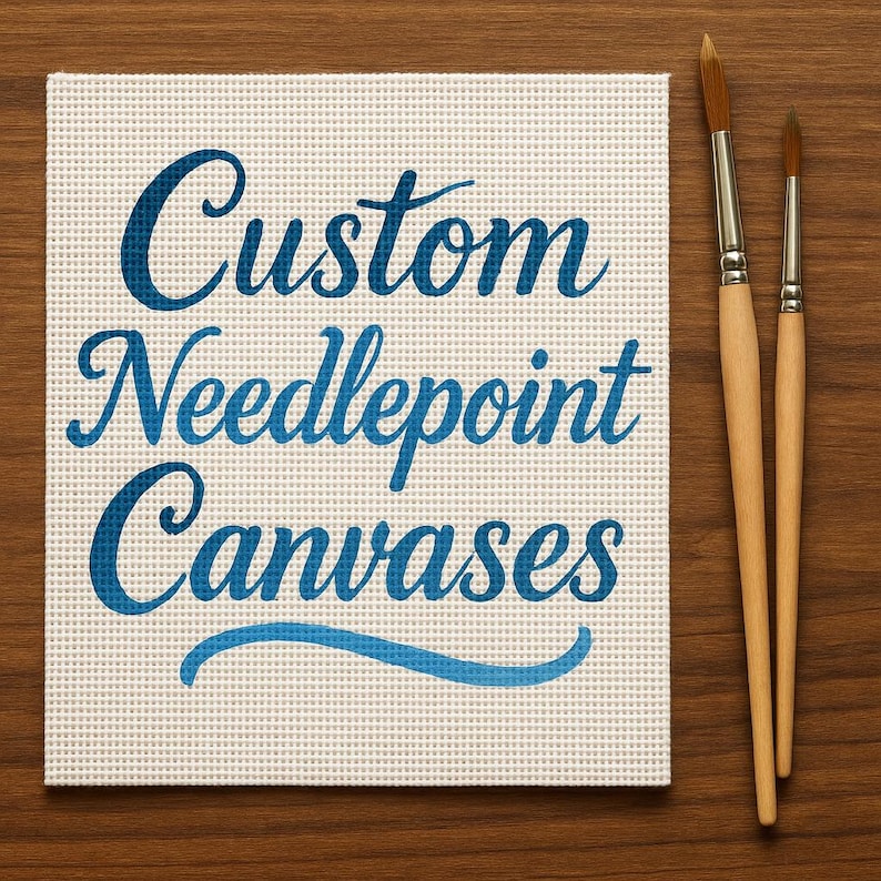 Op de afbeelding: Een vierkant, cr&egrave;mekleurig naaldkunstcanvas met de woorden "Custom Needlepoint Canvases" in blauw script. Twee penselen met houten handvatten liggen op een houten oppervlak.