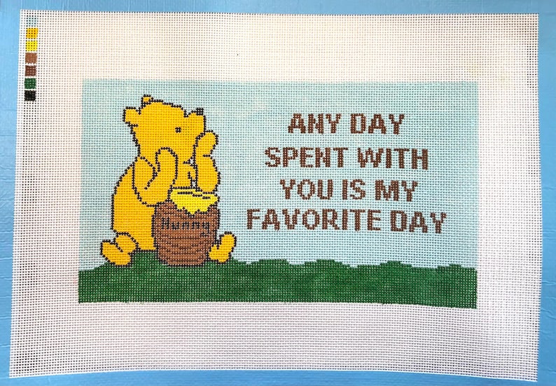 Op de afbeelding: Kruissteek canvas met Winnie de Poeh met een honingpot. De tekst luidt: "ELKE DAG MET JOU IS MIJN FAVORIETE DAG." De achtergrond is lichtblauw met groen gras aan de onderkant. Het canvas staat op een witte achtergrond.