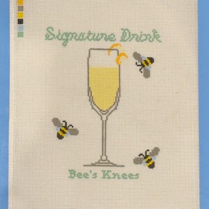 Op de afbeelding: Kruissteekkunst met een champagneglas gevuld met gele vloeistof en een citroenschil. De woorden "Signature Drink" en "Bee's Knees" zijn in lichtgroen. Drie bijen vliegen rond het glas.