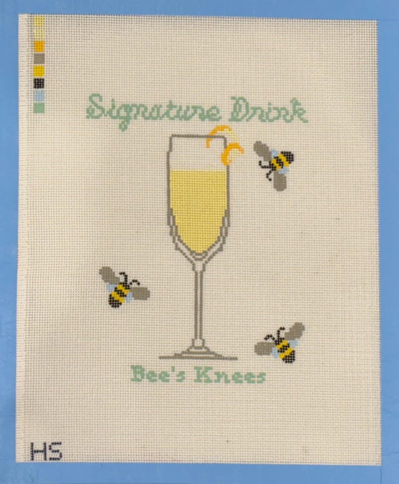 Op de afbeelding: Kruissteekkunst met een champagneglas gevuld met gele vloeistof en een citroenschil. De woorden "Signature Drink" en "Bee's Knees" zijn in lichtgroen. Drie bijen vliegen rond het glas.