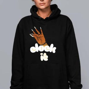 May include: Black hoodie with a graphic of a brown hand with long, white fingernails making a gesture. The words "clock it" are written in a stylized, yellow font below the hand.