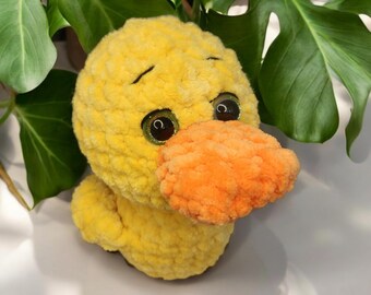 May include: A handmade yellow plush duck with an orange beak and large, glittery eyes. The soft, textured toy is set against a backdrop of green leaves, creating a cheerful and inviting aesthetic.