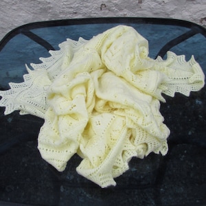 May include: A delicate, pale yellow knitted baby blanket with a lacy border. The blanket is folded and draped on a dark surface.
