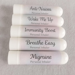 May include: Five white personal inhalers are displayed, each labeled with a different benefit in black script: Anti-Nausea, Wake Me Up, Immunity Boost, Breathe Easy, and Migraine. The inhalers have a rounded shape and are arranged vertically.