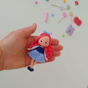 Puede incluir: Una mu&ntilde;eca de ganchillo hecha a mano con cabello rizado rojo, un vestido azul y un lazo azul. La mu&ntilde;eca se sostiene en una mano, con caramelos y n&uacute;meros coloridos en el fondo. La mu&ntilde;eca mide aproximadamente 10 cm de alto.