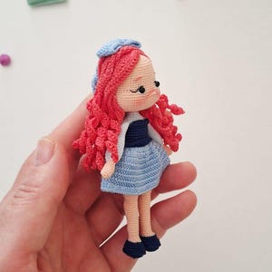 Puede incluir: Mu&ntilde;eca de ganchillo hecha a mano con pelo rizado rojo, que lleva un vestido azul, una parte superior blanca y un sombrero azul. La mu&ntilde;eca tiene zapatos negros y se sostiene en una mano. La mu&ntilde;eca mide aproximadamente 10 cm de altura.