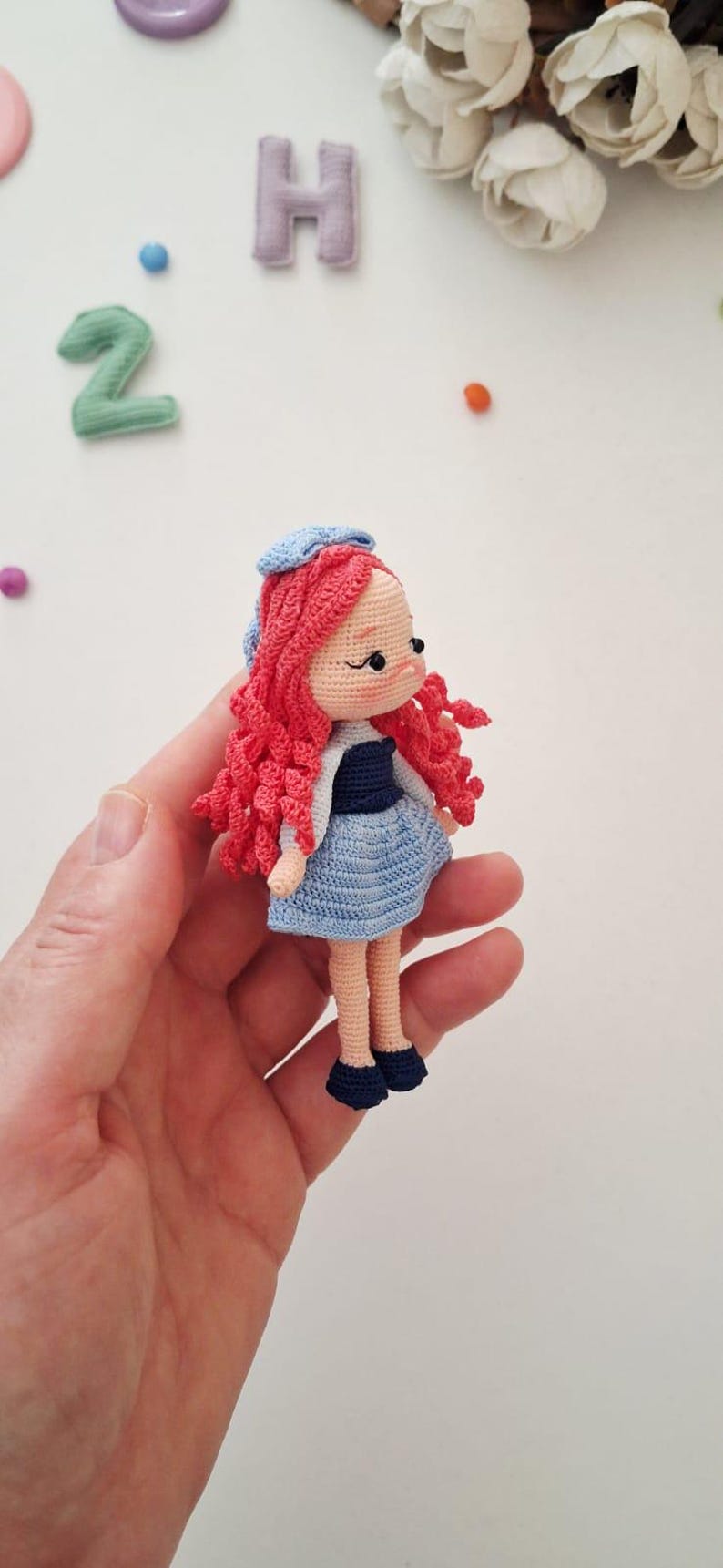Puede incluir: Mu&ntilde;eca de ganchillo hecha a mano con pelo rizado rojo, que lleva un vestido azul, una parte superior blanca y un sombrero azul. La mu&ntilde;eca tiene zapatos negros y se sostiene en una mano. La mu&ntilde;eca mide aproximadamente 10 cm de altura.