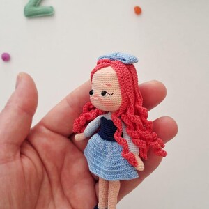 Puede incluir: Mu&ntilde;eca de ganchillo hecha a mano con cabello rizado rojo, un vestido azul y un sombrero azul. La mu&ntilde;eca tiene un tono de piel claro y zapatos negros. La mu&ntilde;eca se sostiene en una mano.