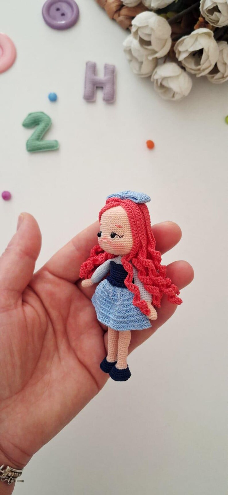 Puede incluir: Mu&ntilde;eca de ganchillo hecha a mano con cabello rizado rojo, un vestido azul y un sombrero azul. La mu&ntilde;eca tiene un tono de piel claro y zapatos negros. La mu&ntilde;eca se sostiene en una mano.