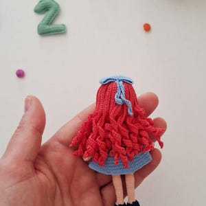 Puede incluir: Una mu&ntilde;eca de ganchillo hecha a mano con cabello rizado rojo, un vestido azul y zapatos negros. La mu&ntilde;eca tiene un lazo azul y se sostiene en una mano. Otros elementos en la imagen incluyen botones y la letra "H".