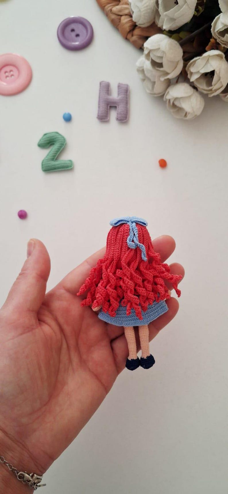 Puede incluir: Una mu&ntilde;eca de ganchillo hecha a mano con cabello rizado rojo, un vestido azul y zapatos negros. La mu&ntilde;eca tiene un lazo azul y se sostiene en una mano. Otros elementos en la imagen incluyen botones y la letra "H".
