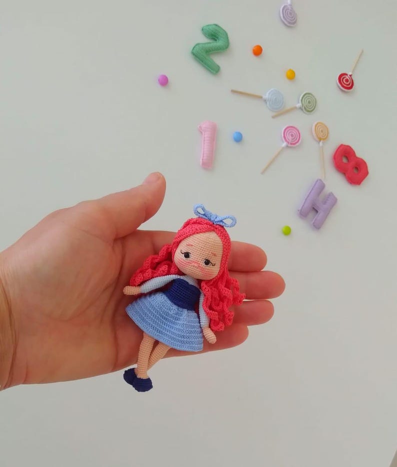 Puede incluir: Una mu&ntilde;eca de ganchillo hecha a mano con cabello rizado rojo, un vestido azul y un lazo azul. La mu&ntilde;eca se sostiene en una mano, con caramelos y n&uacute;meros coloridos en el fondo. La mu&ntilde;eca mide aproximadamente 10 cm de alto.
