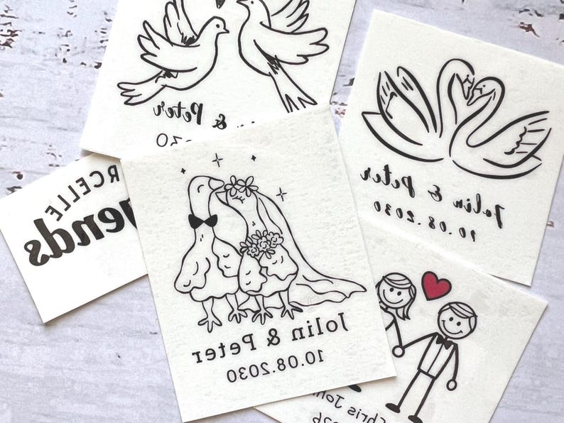 K&ouml;nnte beinhalten: Eine Sammlung wei&szlig;er Papieranh&auml;nger mit schwarzen Strichzeichnungen zum Thema Hochzeit. Zu den Designs geh&ouml;ren Tauben, Schw&auml;ne und Strichm&auml;nnchen sowie Namen und Daten. Das Wort "friends" ist ebenfalls auf einem Anh&auml;nger aufgedruckt.