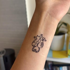 K&ouml;nnte beinhalten: Ein schwarzes Umrisstattoo von zwei Katzen mit Herzen auf einem Handgelenk. Eine Katze hat ein gemustertes Design, die andere Streifen. Die Katzen sind miteinander verschlungen, und kleine Herzen schweben &uuml;ber ihnen. Das Tattoo ist ein einfaches, minimalistisches Design.