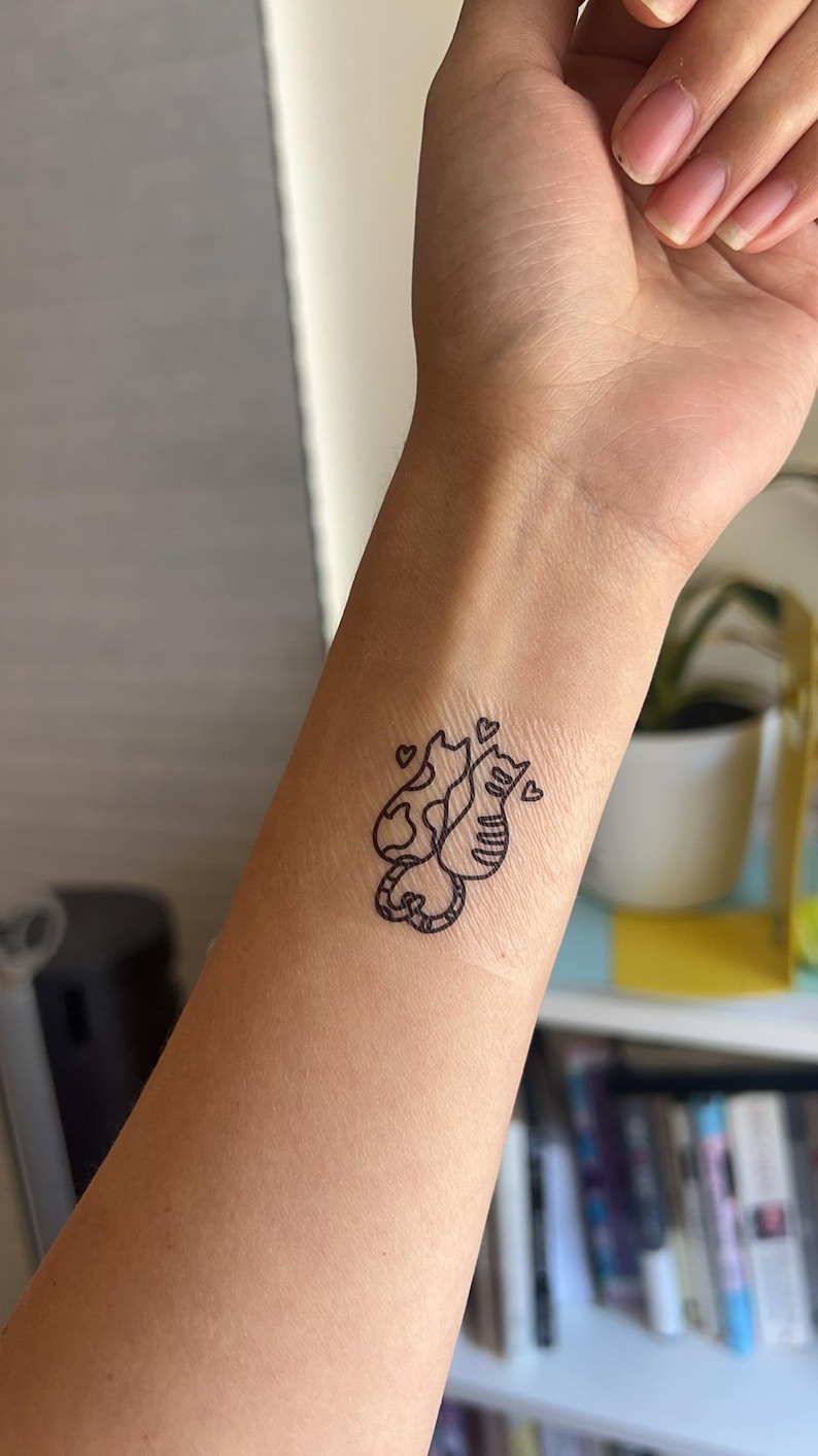 K&ouml;nnte beinhalten: Ein schwarzes Umrisstattoo von zwei Katzen mit Herzen auf einem Handgelenk. Eine Katze hat ein gemustertes Design, die andere Streifen. Die Katzen sind miteinander verschlungen, und kleine Herzen schweben &uuml;ber ihnen. Das Tattoo ist ein einfaches, minimalistisches Design.