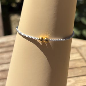 May include: A delicate bracelet featuring a thin, white cord with two small, gold-coloured beads. The bracelet is displayed on a beige arm, with a blurred outdoor background.