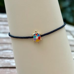 Può includere: Un braccialetto con un ciondolo a forma di tartaruga dorata e un guscio multicolore. Il ciondolo a forma di tartaruga è attaccato a un braccialetto con cordino blu scuro. Il braccialetto è esposto su una superficie chiara.