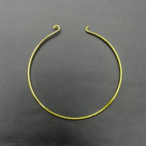 Könnte beinhalten: Eine goldfarbene Halskette mit kreisförmigem Design. Die Halskette hat einen schlichten, eleganten Stil mit einem Hakenverschluss oben. Der Schmuck besteht aus einem dünnen, glatten Metall und ist vor einem dunklen Hintergrund platziert.