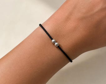 May include: A minimalist bracelet featuring a thin black cord made of twisted thread. The bracelet is adorned with two small, polished silver-toned spherical beads positioned side-by-side at the center of the cord, creating a simple design.