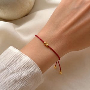 Op de afbeelding: Een dun rood koordarmbandje met twee kleine, gepolijste goudkleurige kralen in het midden van het koord. Het armbandje eindigt met twee bungelende goudkleurige sterbedels, wat zorgt voor een verfijnde en verstelbare afwerking.