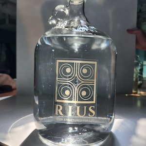 May include: A clear glass water jug filled with water. The jug has a handle and a black cap. A gold label with the word "RIUS" and the text "WATER STRUCTURED FOR LIFE" is on the front.