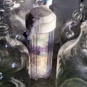 以下が含まれることがあります： 白い上部と青いアクセントが付いた透明な浄水器で、カラフルな石が詰められています。透明なガラス製のガロンジャグがいくつか周囲にあります。 フィルターには「OUT」のラベルと青いバルブが付いています。