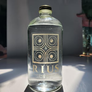 May include: A clear glass bottle with a gold cap and a gold label featuring the word "RIUS" and an abstract design. The bottle is filled with a clear liquid, and the text "WATER STRUCTURED FOR LIFE" is visible.