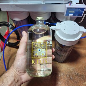 以下が含まれることがあります： 「RIUS WATER STRUCTURED CURE WE」と書かれた金色のラベルが付いた透明なガラス瓶。容量は74ml。手で持っており、背景には浄水設備がある。