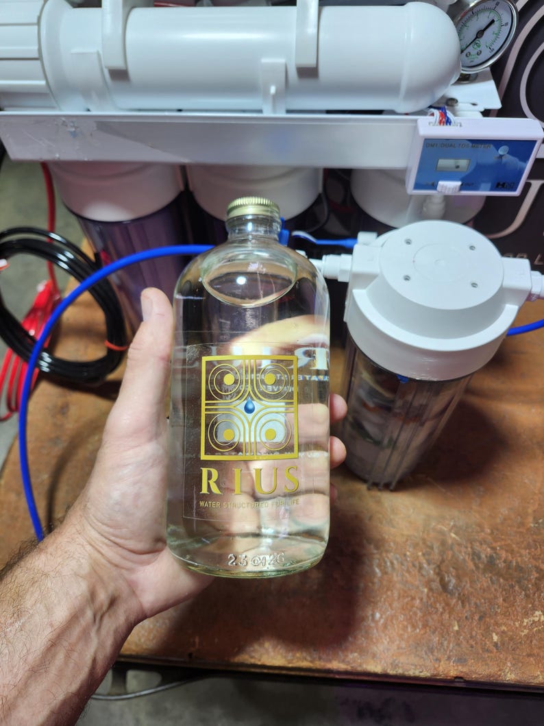以下が含まれることがあります： 「RIUS WATER STRUCTURED CURE WE」と書かれた金色のラベルが付いた透明なガラス瓶。容量は74ml。手で持っており、背景には浄水設備がある。