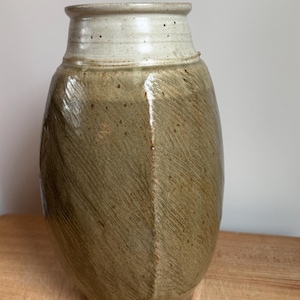 May include: A handmade ceramic vase with a textured, olive-green body and a cream-colored rim. The vase has a unique, asymmetrical shape and a rustic, artisanal aesthetic. The vase is displayed on a wooden surface.