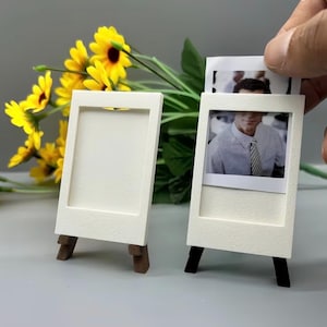 Peut inclure: Deux cadres photo verticaux, rectangulaires et blancs, dotés d'une finition texturée et soutenus par de petits pieds en bois brun foncé. L'un des cadres est vide, tandis que l'autre contient une petite photo d'une personne en chemise.