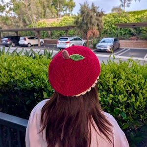 Puede incluir: Boina roja de ganchillo con un diseño decorativo de manzana en la parte superior. La manzana tiene un tallo marrón y una hoja verde. La boina tiene un borde festoneado de color blanquecino. El fondo incluye follaje verde y coches aparcados.