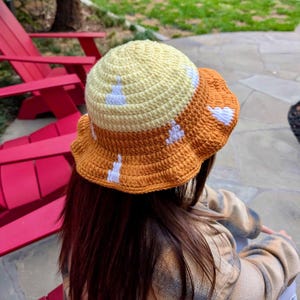 May include: A handmade crochet bucket hat in yellow and orange. The hat features white bunny designs and a scalloped brim. The hat is worn by a person with long brown hair.