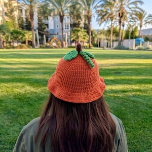 May include: An orange crochet bucket hat with green leaves and a brown stem, resembling a pumpkin. The hat is worn outdoors in a park setting. The background features palm trees and buildings.