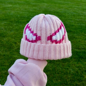 May include: A light pink knitted beanie with a ribbed brim. The hat features a design of white and magenta shapes on the front. The hat is held up by a person wearing a light pink sweatshirt. The background is green grass.
