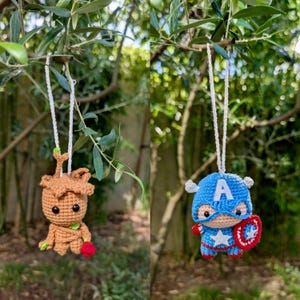 Puede incluir: Dos adornos de ganchillo colgando de una rama de árbol. Uno es una figura marrón con una rosa roja, el otro es el Capitán América azul con un escudo. Ambos están suspendidos por cuerdas blancas.