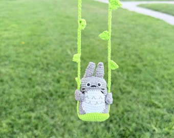 May include: A gray and white crocheted figure with tall ears and a round belly, sitting on a bright green crocheted swing. The swing is suspended by two long, thin green crocheted vines featuring small, pointed green leaves attached to the sides.